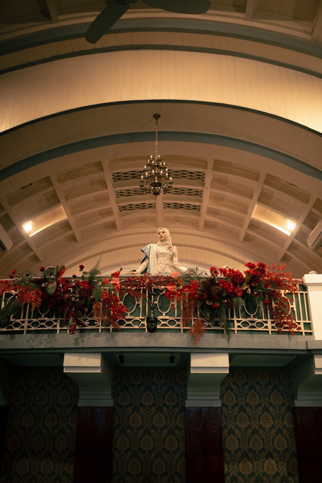 Graduate standing on a symmetrical balcony framed by red flowers and a vaulted ceiling.